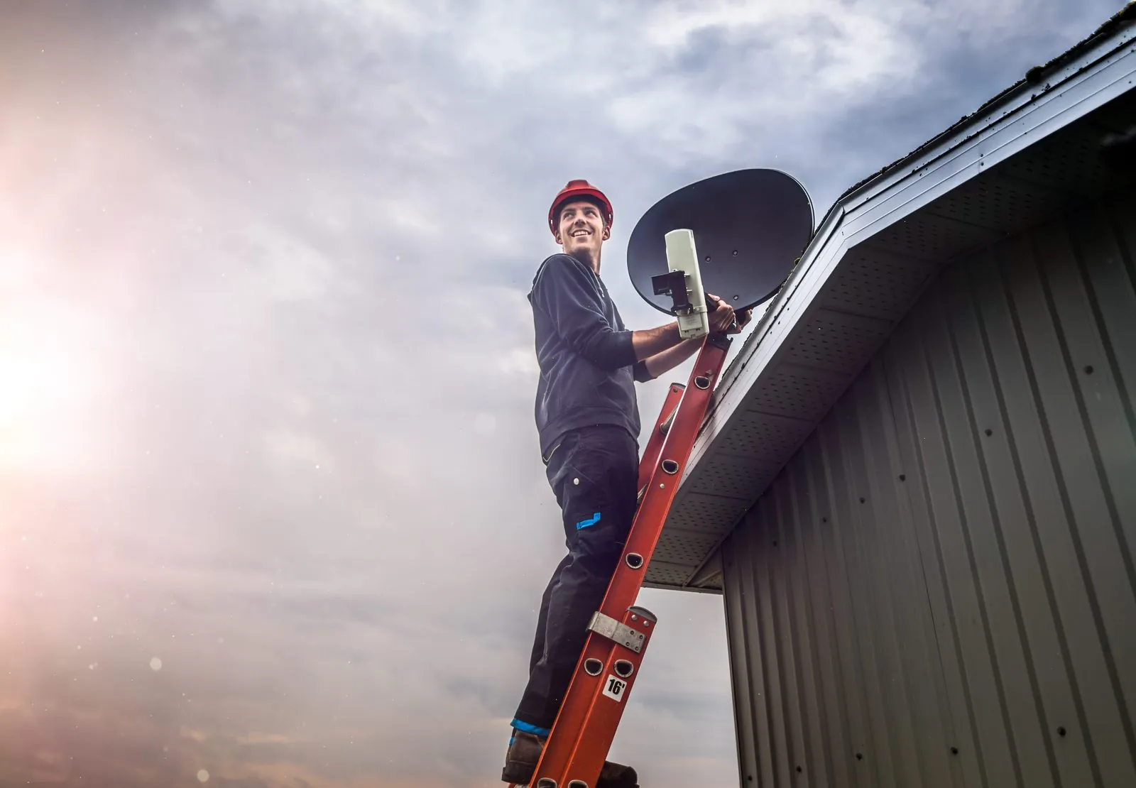Technician on a ladder installing a satellite dish at the edge of a roof